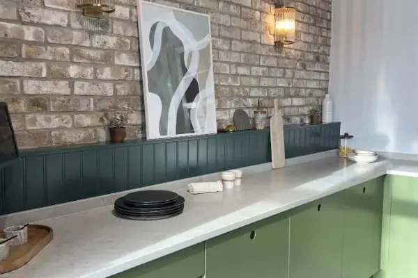 An exposed brick wall with a white kitchen countertop and sage green cabinets.