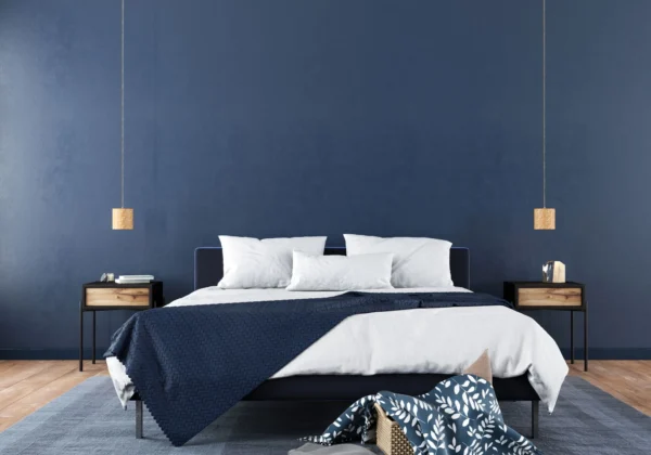 Stylish bedroom interior with bold dark blue feature wall, lighter blue rug and dark blue throw. There are black bedside tables with wooden drawers, and the floor is also wooden.