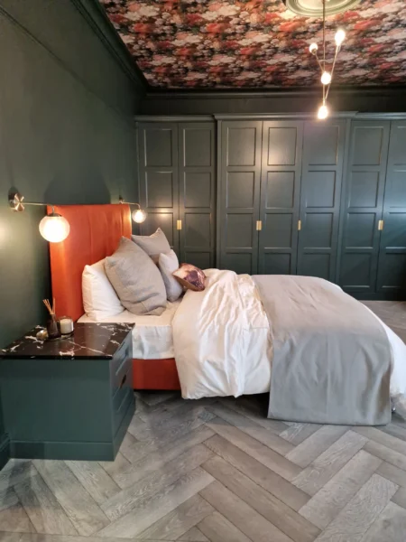 A bedroom with dark green walls and a floral ceiling. There are built-in dark green wardrobes, an orange headboard, and a herringbone floor.