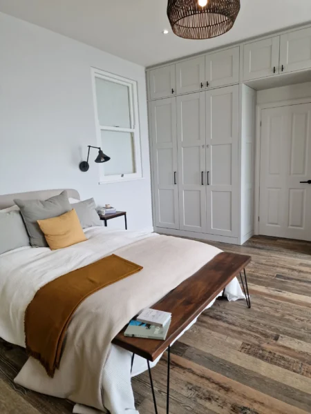 A light-coloured bedroom with built-in white wardrobes and a bed with a wooden bench at the foot.