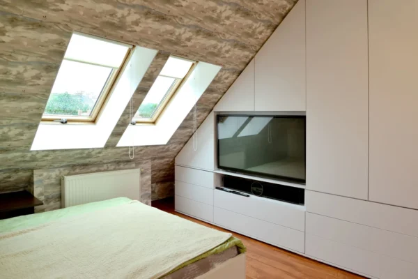 A modern attic bedroom with a sloped ceiling, skylight windows, and a built-in white wardrobe with a TV.