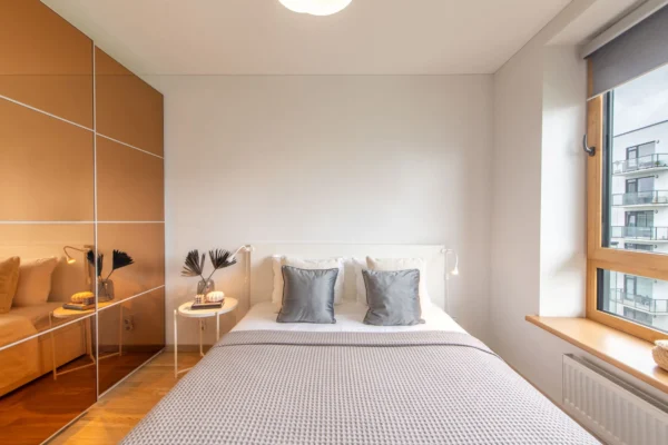 A minimalist bedroom with white walls, a white bed, and a mirrored wardrobe reflecting the room.