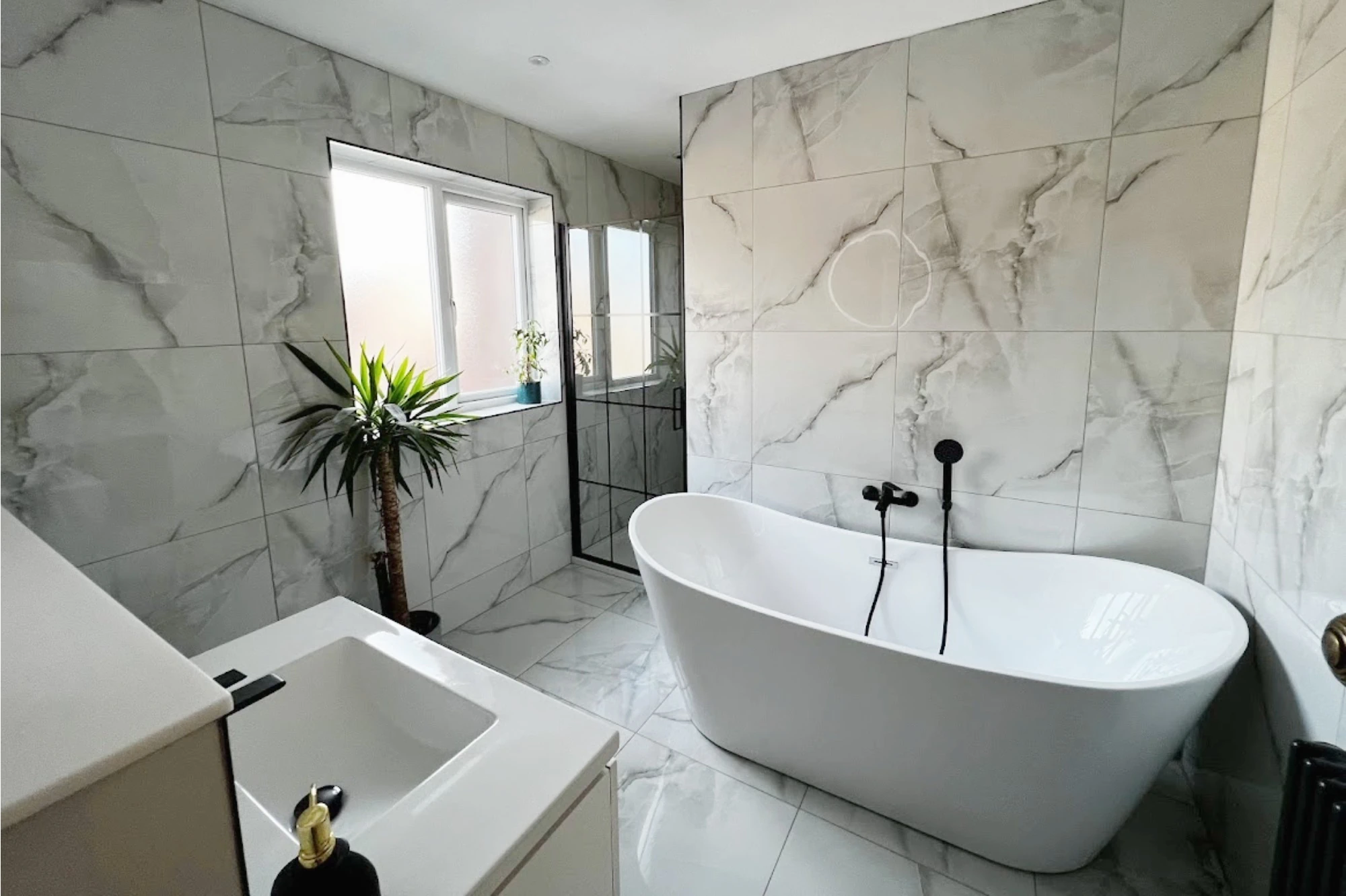 A stylish bathroom designed by Premier Crafts with walls and floor tiled in white marble-effect tile. A large freestanding bathtub with black fixtures is positioned in the centre, and a modern vanity with a rectangular basin is in the foreground. 