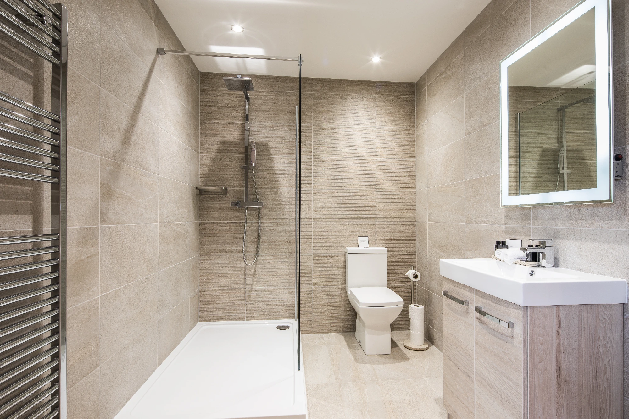 A compact modern bathroom with light-coloured tile walls, a walk-in shower with a glass panel, a white toilet, and a small vanity with an illuminated mirror. 