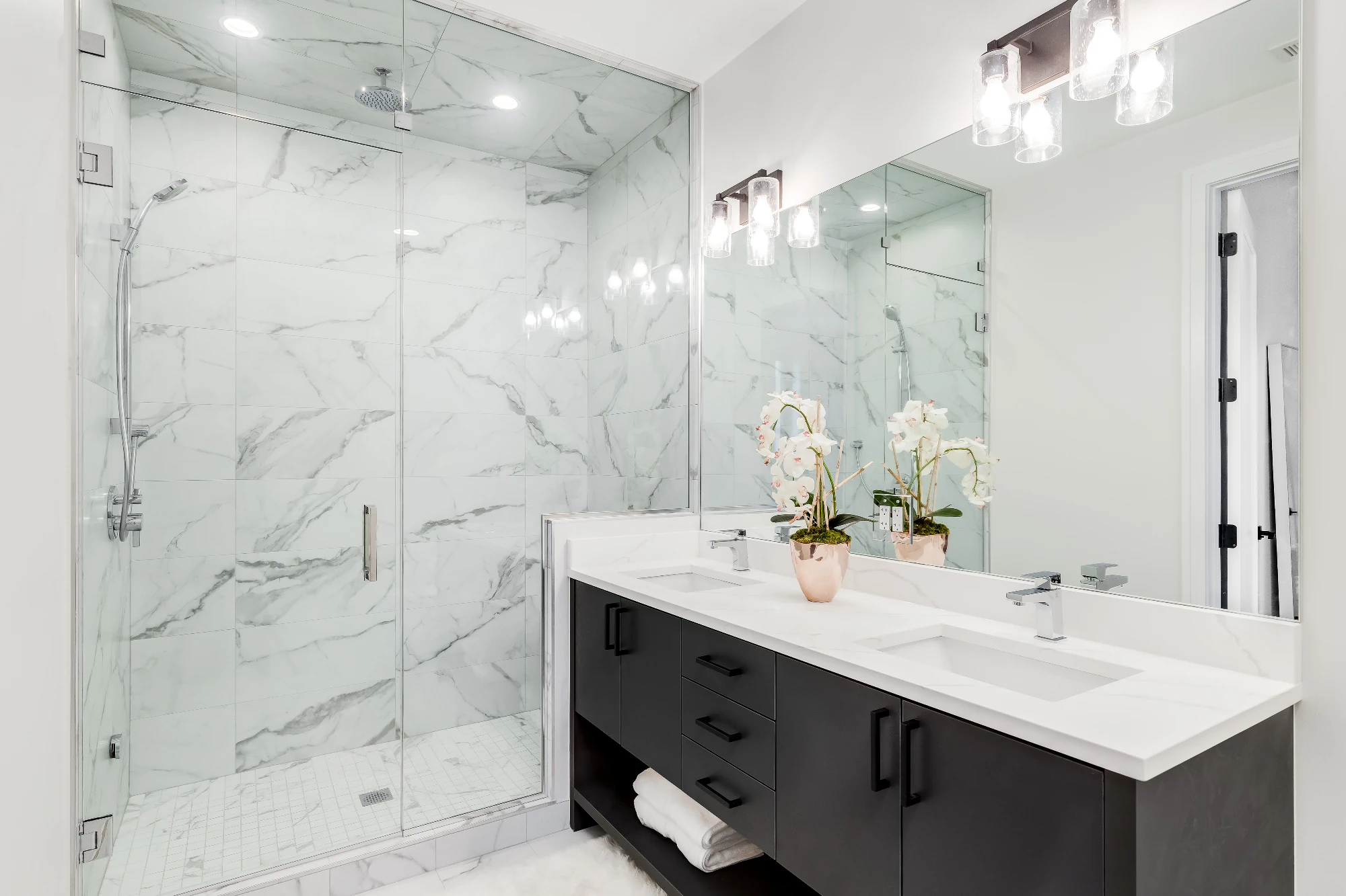 A clean and modern bathroom with a double vanity and a glass-enclosed walk-in shower with white marble walls and a subway tile floor. 