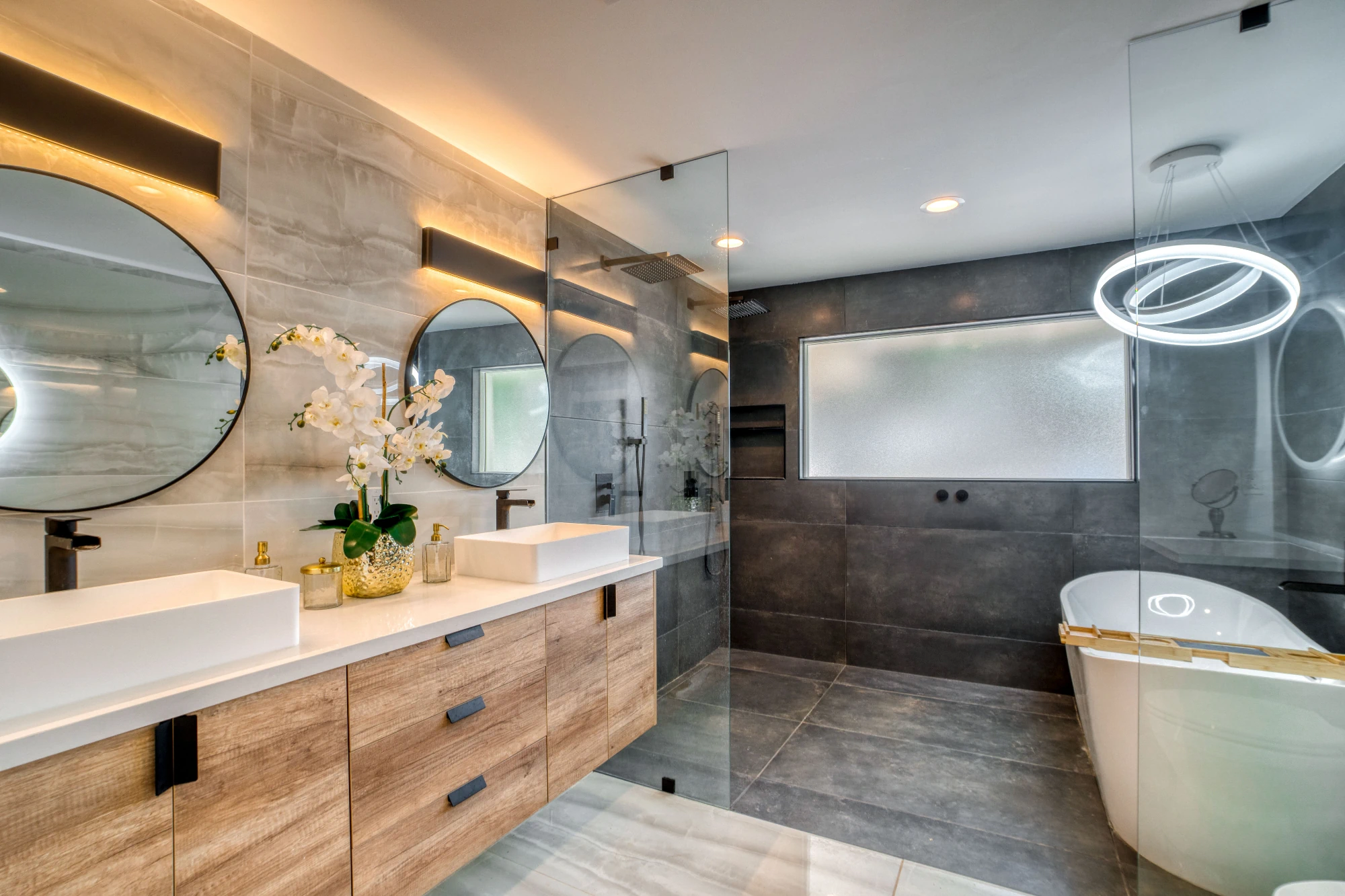 A stylish modern bathroom with a light wood double vanity, two round mirrors with backlighting, a walk-in shower with a glass panel, and a white freestanding bathtub with a contemporary ring chandelier. 