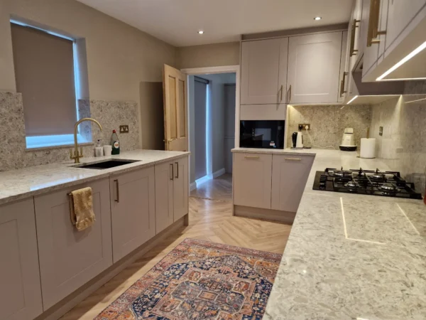 A modern galley kitchen with light grey cabinets, speckled quartz countertops, and a herringbone wood floor.