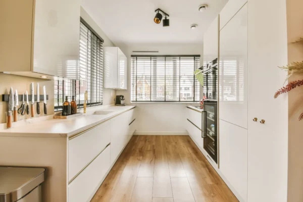 A modern galley kitchen with high-gloss white cabinets, wood flooring, and large windows with black blinds.