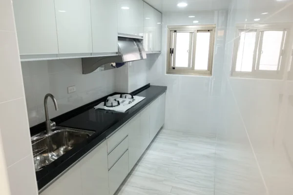 A modern, white galley kitchen featuring high-gloss cabinetry, white tiled walls, and a black countertop with an integrated gas hob and stainless steel sink.