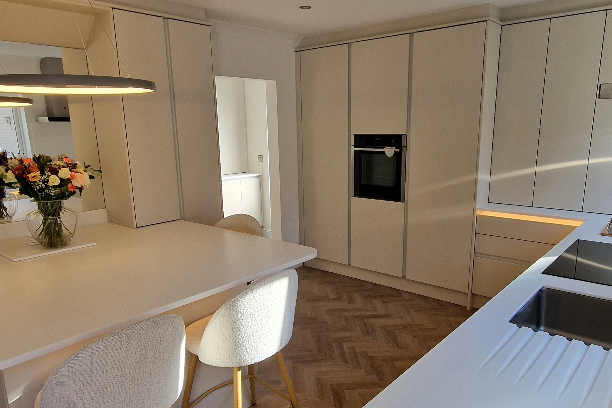 A modern kitchen with sleek white cabinetry, peninsula, and textured chairs bathed in sunlight.