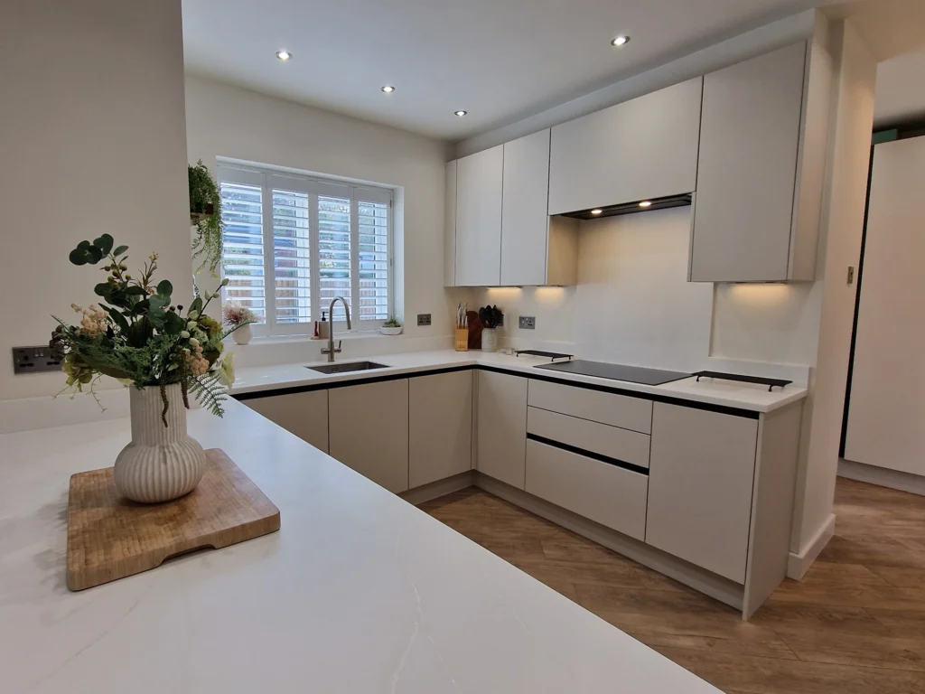 A handleless white kitchen featuring under-cabinet lighting, a built-in induction hob, and a large white island with a decorative floral arrangement.