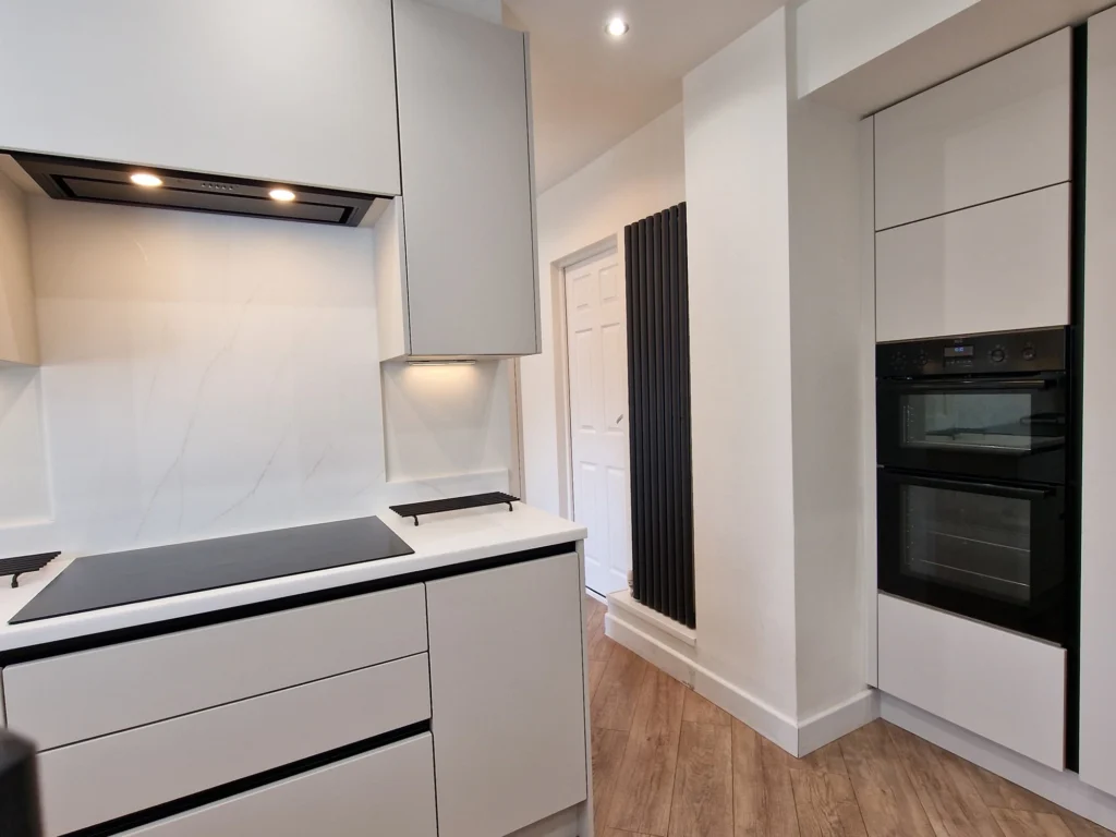 A modern white kitchen featuring handleless cabinets, a built-in black double oven, and a white marble-patterned backsplash.