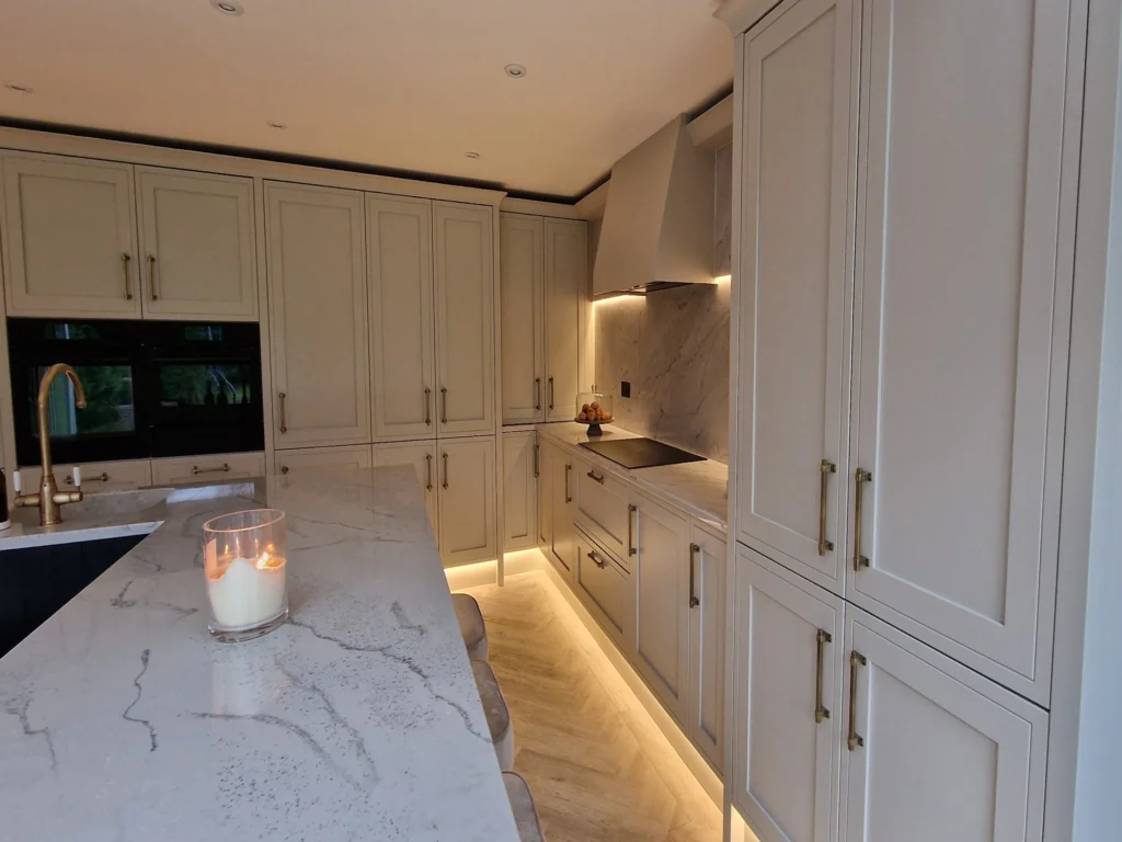 A traditional-style kitchen featuring light grey Shaker cabinets with gold hardware, a marble backsplash, and warm under-cabinet lighting.