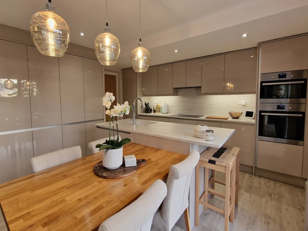 A contemporary, high-gloss kitchen featuring a large central island, three elegant glass pendant lights, and an integrated wooden dining table.