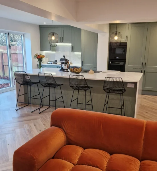 A sage green kitchen with white ceilings, a white kitchen island with 4 high seats, and an orange sofa.