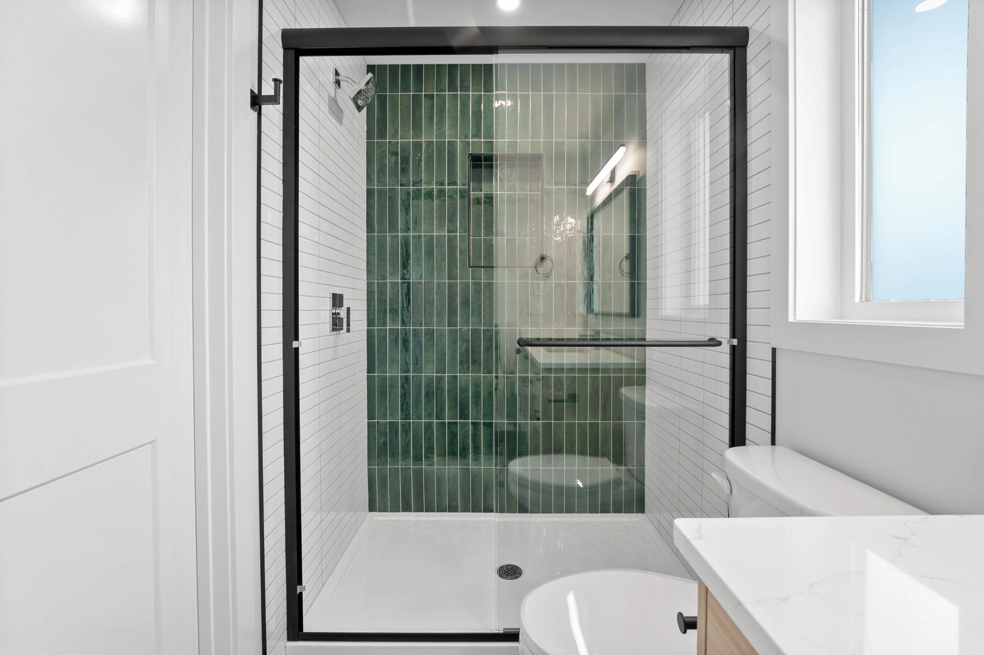 White compact bathroom with wall-to-wall glass door shower with black detailing and green tiles.