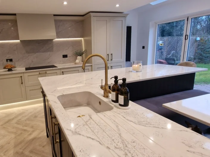 Contemporary shaker-style kitchen featuring a large marble-veined island with a brass faucet, off-white cabinetry with gold handles, under-cabinet lighting, and light herringbone wood flooring.