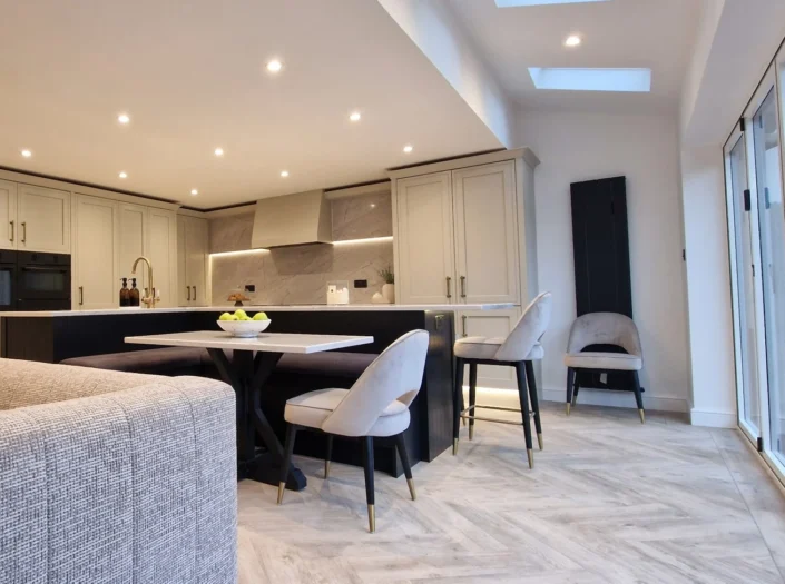Open-concept kitchen and living area with a large marble island, built-in banquette seating, and floor-to-ceiling off-white cabinetry under bright recessed lighting.