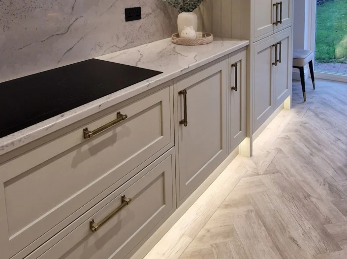 Close-up of off-white shaker-style kitchen drawers featuring antique brass handles and warm LED plinth lighting reflecting on a light oak herringbone floor.