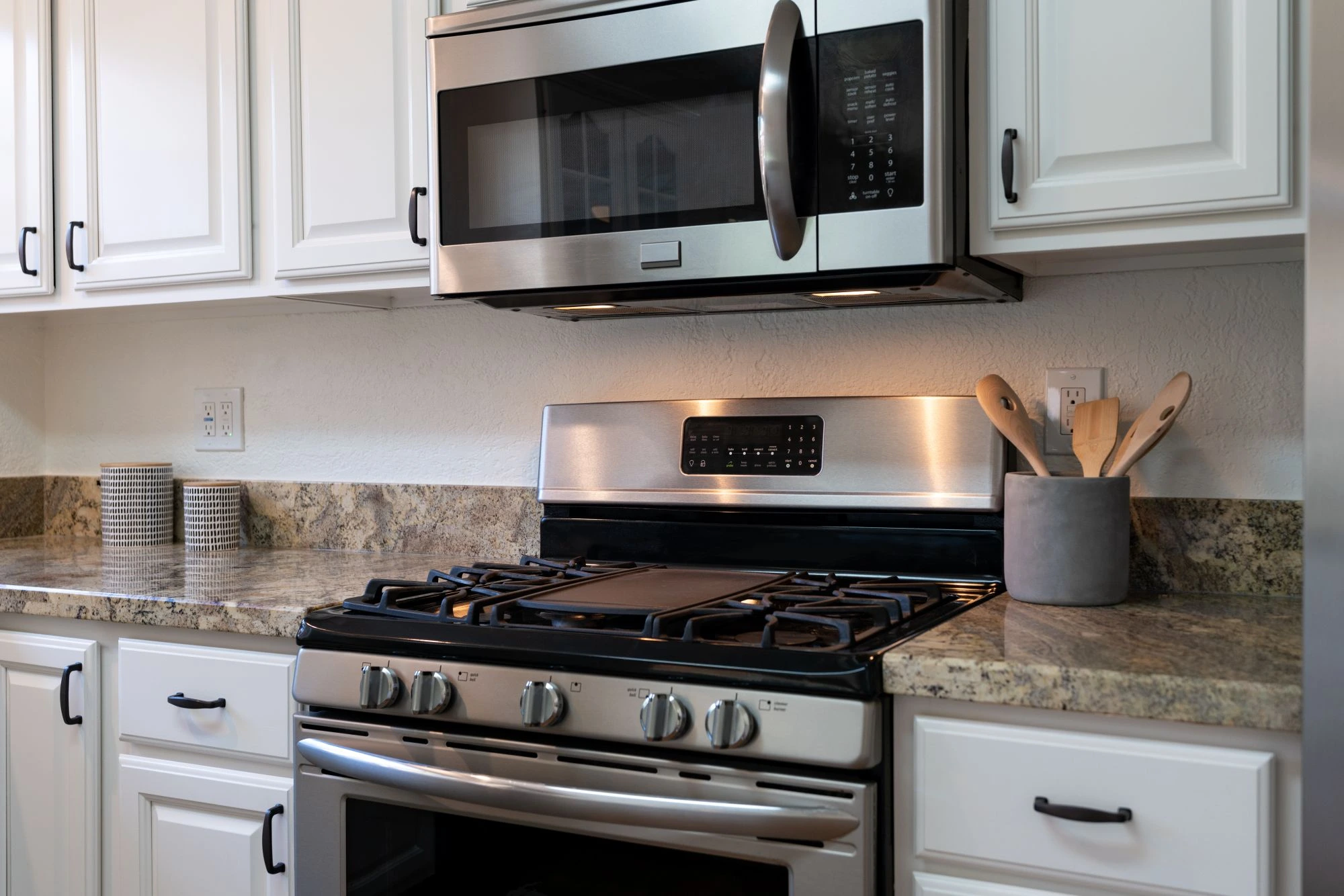 Stainless steel gas range and over-the-range microwave built into white kitchen cabinets with granite countertops.