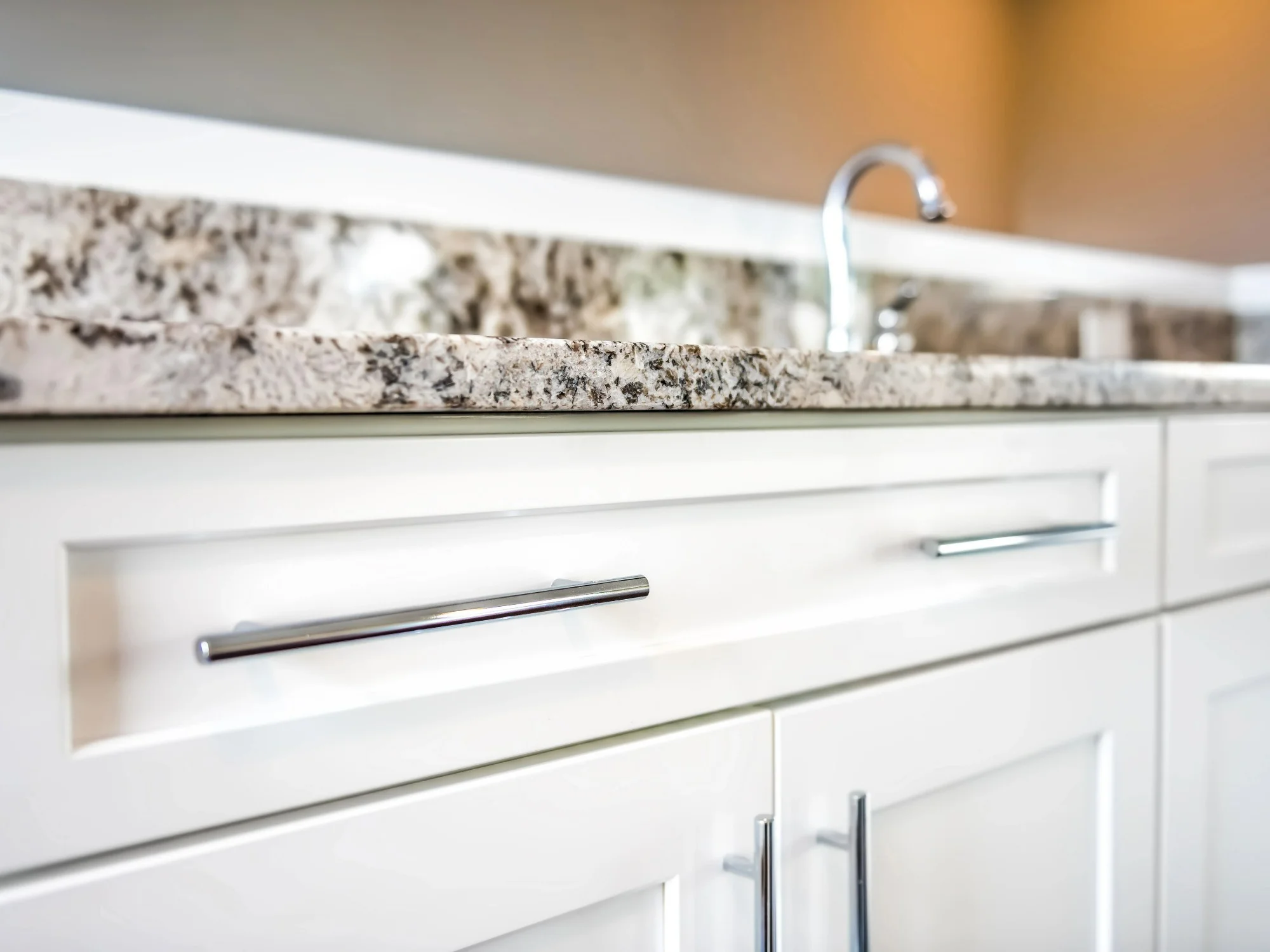 Close up of white kitchen cabinets with silver handles and a textured granite worktop.